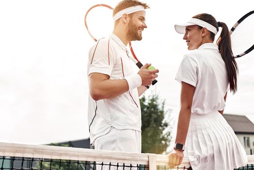 Beautiful woman and handsome man are going to play tennis. Woman in white polo and skirt looks ar her partner. Man in white sportswear with the racket and the ball in his hands looks at her