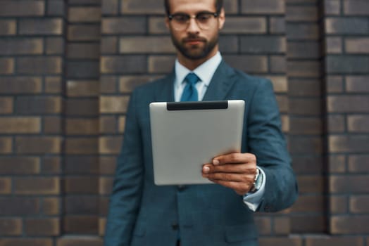 Modern businessman. Portrait of handsome bearded businessman in eyeglasses working with touchpad while standing against brick wall. Digital concept. Business concept