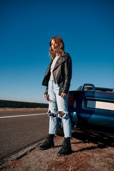 Young woman near roofless car and smiling outdoors on sunny day near the empty road. No people, sportive blue car