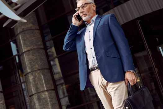 Successful businessman. Serious senior man in casual suit talking on the phone while walking outdoors