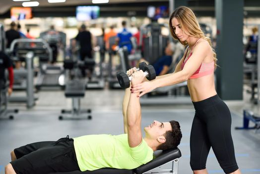 Female personal trainer helping a young man lift weights while working out in a gym