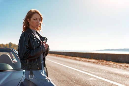 Young woman near roofless car and smiling outdoors on sunny day near the empty road. No people, sportive black car