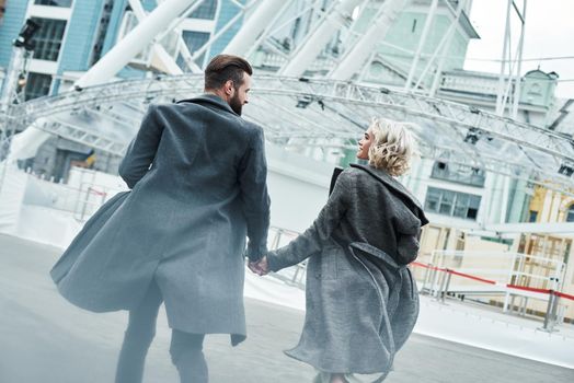 Romantic date outdoors. Young couple running at entertainment park holding hands back view looking at each other smiling happy