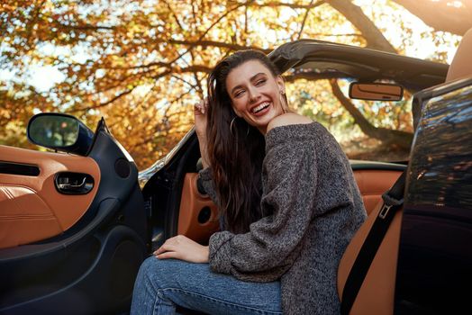 Sexy young woman in jeans and in a grey sweater sits in front seat of a cabriolet. Autumn season.