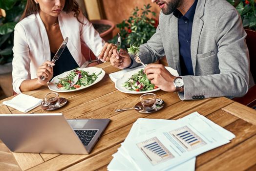 Business lunch. Man and woman sitting at table at restaurant eating healthy fresh salad discussing project close-up