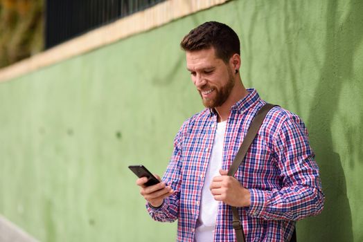 Young man looking at his smart phone and smiling in urban background. Guy wearing casual clothes. Lifestyle concept.