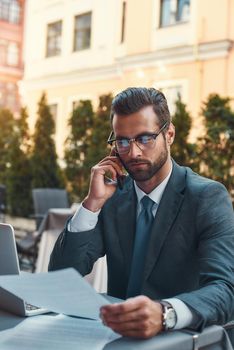 I am working everywhere. Portrait of handsome bearded businessman in eyeglasses talking by phone with client and looking at documents while sitting in restaurant outdoors. Digital concept. Business concept