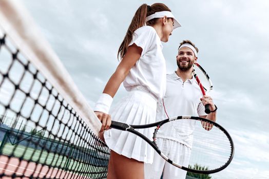 Lovers in white sportswear with rackets in their hands look at each other while going to play tennis on the court