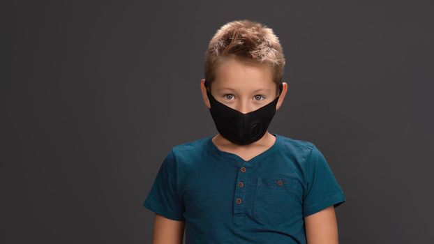 School boy in black reusable face mask wearing dark blue button neck t-shirt surprised look at camera while standing in studio. Isolated on black background. Kids fashion concept.