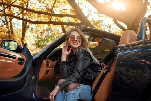 Sexy young woman in jeans and in a leather jacket sits in front seat of a cabriolet. Autumn season