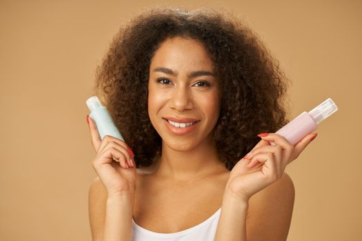 Excited young mixed race woman smiling at camera, holding two bottles with beauty product while posing isolated over beige background. Skincare concept. Horizontal shot