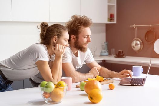 Leaned on kitchen table couple watching romantic movie or streaming social media video. Beautiful young couple talking on video call using laptop. Young couple in kitchen at new home.