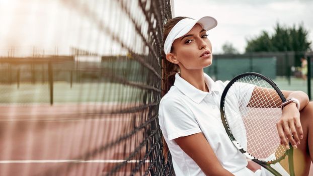 Young female tennis player sitting on a court after match and holding a racket, looking away. Healthy lifestyle. Horizontal shot