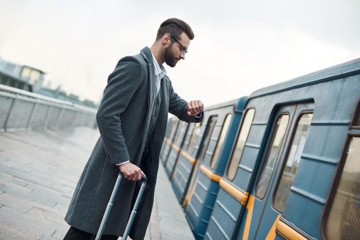 Business trip. Young businessman standing near railway with luggage checking time on watch waiting for train