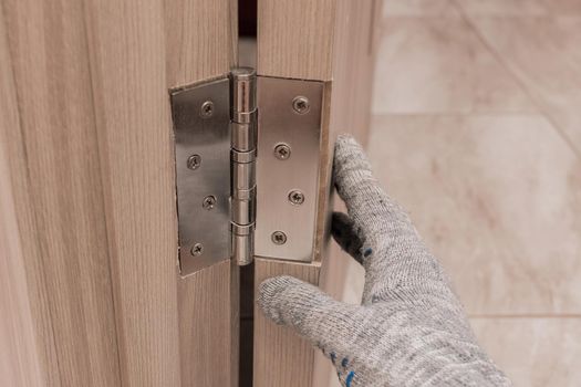 The hand of a working man in a construction glove touches and examines a wooden door opening with a door hinge, close-up. Installation or adjustment of metal parts.