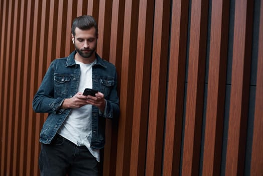 Freestyle. Young man standing leaning on wooden wall isolated browsing internet on smartphone concentrated