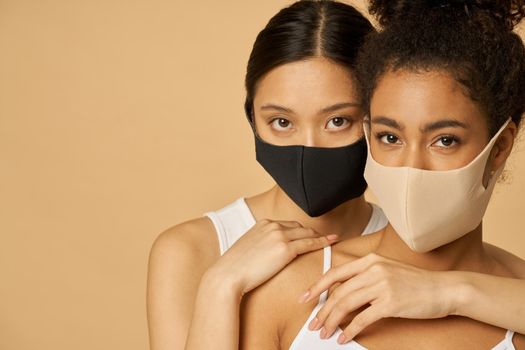 Two attractive young women, female friends wearing protective facial masks while posing together isolated over beige background. Safety, pandemic concept