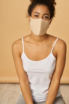 Studio shot of attractive young mixed race woman wearing protective facial mask looking at camera while sitting over beige background. Safety, pandemic concept