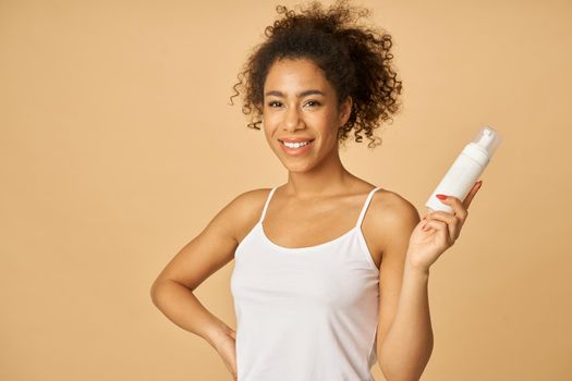 Attractive young woman smiling at camera, holding a bottle of foam facial cleanser, posing isolated over beige background. Beauty products and skin care concept