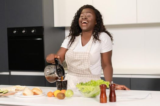 African Woman in Apron Pours Black Coffee from a Glass Coffee Maker into a Mug. Woman Enjoy Cooking her Breakfast. Close-up. Kitchen Background