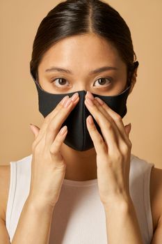 Studio portrait of young woman putting on black facial mask, looking at camera while posing isolated over beige background. Safety, pandemic concept