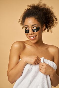 Young woman wrapped in white towel after shower looking playful, smiling and posing with applied black under eye patches isolated over beige background. Bathroom routine concept