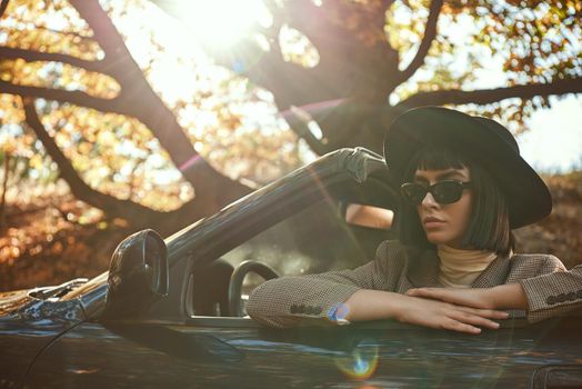 Sexy lady in the roofless car. Stylish woman is wearing sunglasses and black hat. She had fashion haircut. Autumn season.