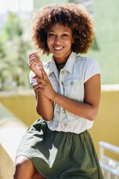 Young black woman, afro hairstyle, smiling. Girl, model of fashion, wearing casual clothes in urban background. Female with skirt and denim vest.