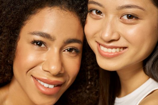 Face closeup of two beautiful happy mixed race young women with perfect glowing skin smiling at camera while posing together. Skincare, diversity concept. Selective focus. Horizontal shot