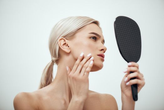 Perfection. Profile portrait of young attractive woman touching her face and looking in small mirror while standing against grey background. Skin care. Beauty concept