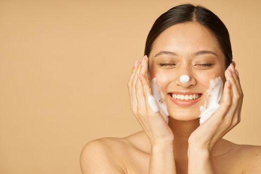 Studio portrait of pleased young woman smiling with eyes closed while applying gentle foam facial cleanser isolated over beige background. Beauty products and skin care concept. Front view