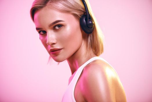 Music in my soul. Close up of cute blonde woman in headphones listening music and looking at camera while standing against pink background. Sport. Music concept