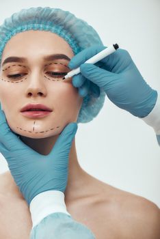 Beautiful patient. Young woman in blue medical hat waiting for facial surgery while plastic surgeon in blue gloves drawing dashed lines on her face. Beauty concept. Facelift. Plastic surgery
