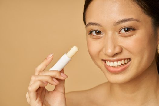 Face closeup of adorable young woman with beautiful smile looking at camera and holding lip balm, posing isolated over beige background. Beauty treatment concept