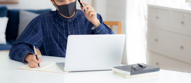 Young asian business man in face mask talking smartphone and work on laptop computer for protect covid-19, businessman quarantine work from home and writing notebook, social distancing and stay home.