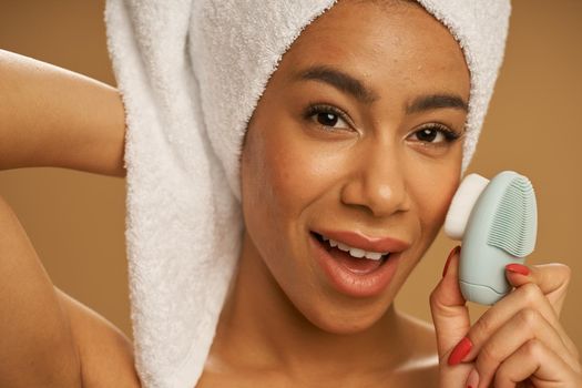 Face closeup of playful young woman looking at camera while using silicone facial cleansing brush, posing isolated over beige background. Skincare concept