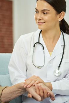 Caring nurse holding the hands of elderly male patient and sitting on the couch at his home