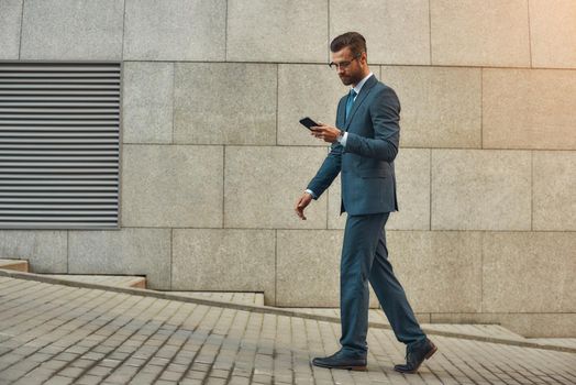 Business message. Side view of handsome businessman in formal wear using smart phone while walking outdoors. Business concept. Digital concept