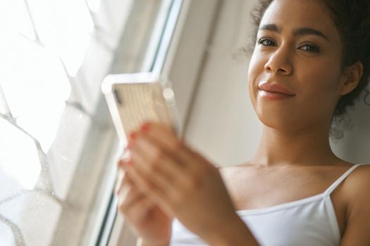 Portrait of young mixed race woman looking at camera, using smartphone, feeling cozy while sitting by the window at home. Lifestyle, leisure concept