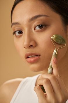 Close up portrait of beautiful young woman looking at camera and using jade roller for massaging her face, posing isolated over beige background. Skincare concept. Vertical shot