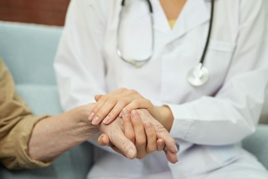 Close up of caring female doctor holding the hands of elderly male patient and sitting on the couch at his home