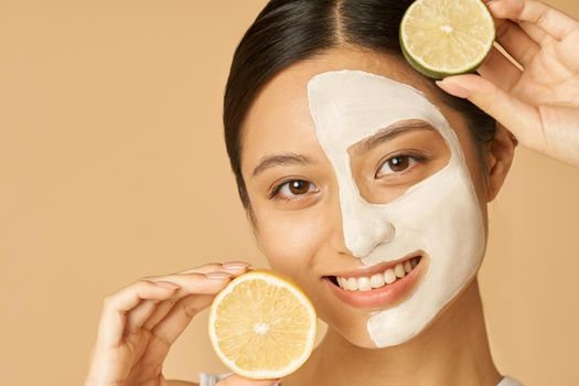 Beauty portrait of young woman with facial mask applied on half of her face holding slice of lime and lemon, posing isolated over beige background. Skincare, natural cosmetics concept