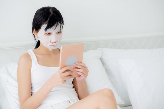 Beautiful young asian woman with sheet facial mask sitting watching tablet computer on bed at bedroom, beauty girl applying face mask for skincare of wrinkle at home, skin care, health and wellness.