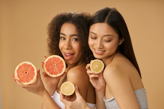 Portrait of two lovely mixed race young women holding grapefruit and lemon cut in half while posing isolated over beige background. Health and beauty concept