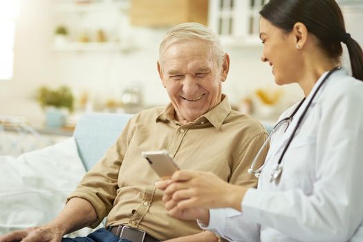 Happy female doctor holding smartphone while talking to senior man at his home