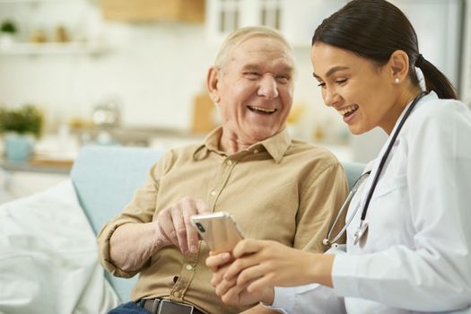 Smiling female doctor holding smartphone while talking to aged man at his home