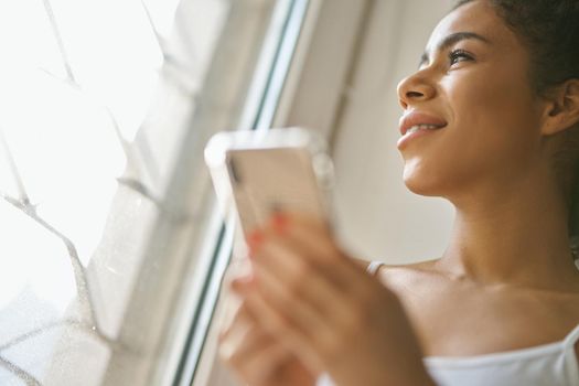 Portrait of young mixed race woman looking out the window, holding smartphone while sitting by the window at home. Lifestyle, leisure concept