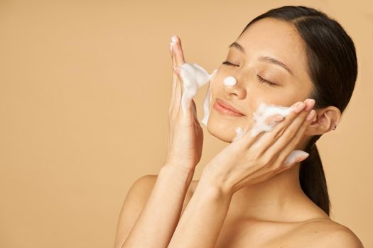 Beauty portrait of happy young woman smiling with eyes closed while applying gentle foam facial cleanser isolated over beige background. Beauty products and skin care concept