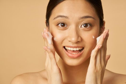 Studio portrait of excited young woman smiling at camera while applying gentle foam facial cleanser isolated over beige background. Beauty products and skin care concept. Front view