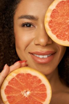 Face closeup of lovely mixed race young woman smiling at camera, posing with grapefruit cut in half. Health and beauty concept. Front view